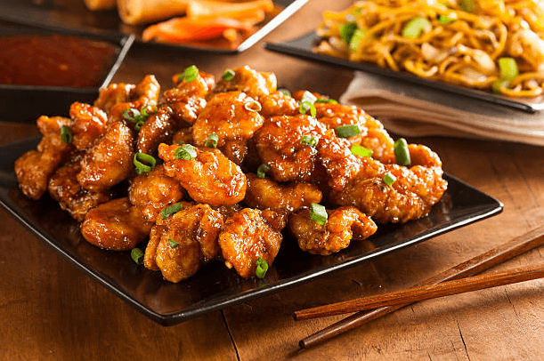 Golden crispy orange chicken coated in a glossy orange marmalade sauce, garnished with sesame seeds and green onions, served on a white plate.