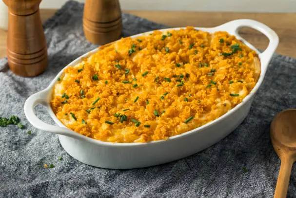 Close-up of a freshly baked old-fashioned scalloped corn dish with golden, crunchy breadcrumbs and creamy custard. Visible corn kernels and ingredients like fresh corn, milk, eggs, butter, and breadcrumbs are subtly displayed around the dish in a rustic setup, with warm natural lighting highlighting the textures and colors.