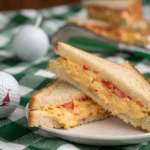 Classic pimento cheese sandwich served on white bread, wrapped in green wax paper, with a golf-themed backdrop at Augusta National Golf Club.