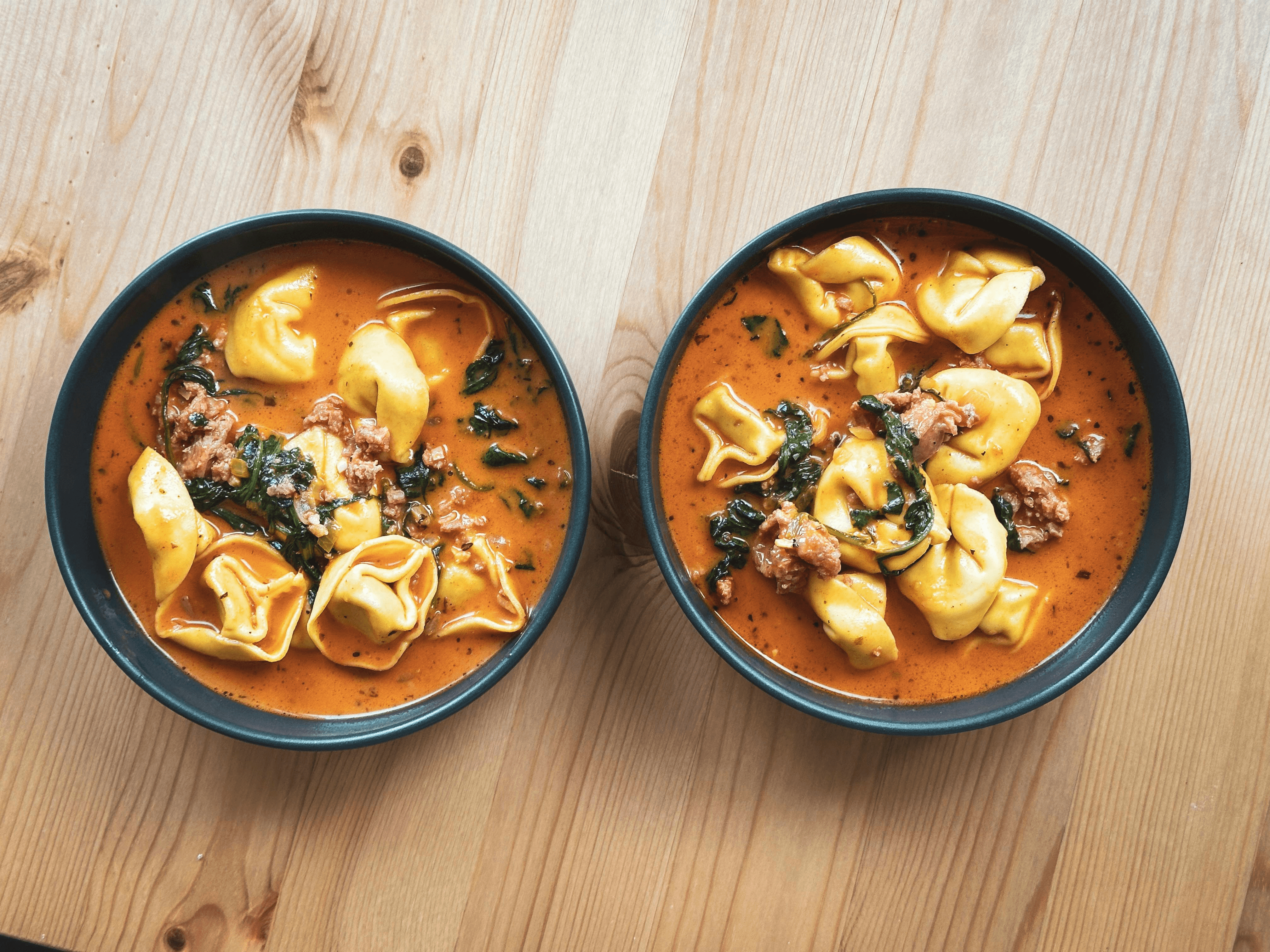 Steaming bowl of tomato tortellini soup garnished with fresh basil and Parmesan cheese, served with a wooden spoon and crusty bread in a sunlit kitchen setting.